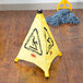 A Rubbermaid yellow wet floor cone with black "Caution" text sits on a wood floor next to a mop.