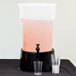 A Carlisle clear plastic beverage dispenser with a pink drink and black base on a table with clear plastic cups.