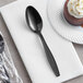 A Visions black plastic teaspoon on a white napkin next to a chocolate cake.