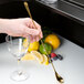 A hand holding a gold Barfly Japanese style bar spoon next to a glass with a lemon slice.