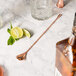 A copper plated Barfly classic bar spoon on a marble surface with a bottle of alcohol and a glass of liquor.