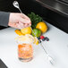A hand holding a Barfly stainless steel bar spoon with fork end next to a glass of juice with ice and lemon slices.