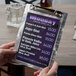 A hand holding a Menu Solutions Alumitique aluminum menu board with a swirl finish.