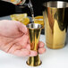 A hand using a Barfly gold-plated Japanese style jigger to pour liquid into a gold cup.