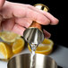 A person using a Barfly gold-plated Japanese style jigger to pour liquid into a metal cup with lemons.