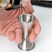 A person using a Barfly stainless steel Japanese style jigger to pour liquid into a small metal cup.