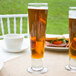 Two Libbey footed pilsner glasses filled with beer on a table with a sandwich.