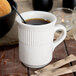 An Ivory embossed rim china mug filled with coffee and a straw on a table with bread and candles.