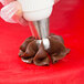 A hand using an Ateco Russian ball tip piping tip to pipe chocolate over cookies on a red surface.