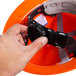 A hand holding a black strap on a Cordova Duo Safety orange hard hat.