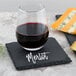 A glass of wine on a slate coaster next to a cheese plate with the word "merlot" written in chalk.