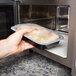 A hand placing a Pactiv Newspring black plastic rectangular container of food into a microwave.