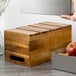 A hand holding a Tablecraft acacia wood serving crate with red apples inside.