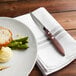 A Walco stainless steel steak knife on a napkin next to a plate of food with mashed potatoes and asparagus.