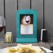 A Menu Solutions sky blue wood menu frame on a table with a glass of wine and a plate of chips.