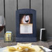 A Menu Solutions denim wood menu tent on a table with a glass of wine.