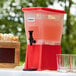 A red and white Choice 3 gallon beverage dispenser with glasses on a table.