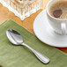 An Acopa stainless steel demitasse spoon on a green napkin next to a cup of coffee on a saucer.