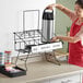 A woman in a red apron places a Choice black wire 3-compartment airpot rack on a counter.