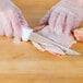 A person in gloves uses a Choice 6" Wide Stiff Boning Knife to cut up chicken on a wooden table.