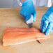 A person wearing blue gloves using a Choice curved stiff boning knife to cut a piece of salmon.