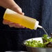 A hand using a Choice clear squeeze bottle to pour yellow mustard onto a bowl of green salad.