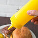 A hand holding a yellow Choice wide mouth squeeze bottle pouring mustard onto a bun.