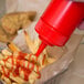 A hand pouring ketchup from a red Choice wide mouth squeeze bottle onto a basket of french fries.