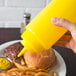 A hand holding a yellow Choice wide mouth squeeze bottle over a plate of fries and hamburgers.