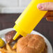 A hand holding a yellow Choice squeeze bottle over a burger and fries.