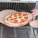 An American Metalcraft round pizza peel with a person's hands placing a pizza with cheese and pepperoni into an oven.