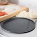 A Matfer Bourgeat black carbon steel pizza pan on a counter with pizza dough and ingredients.