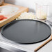 A Matfer Bourgeat black carbon steel pizza pan on a counter with pizza dough and cheese.
