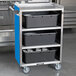 A Lakeside stainless steel utility cart with a blue enclosed base and four metal shelves holding black containers.