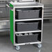 A Lakeside stainless steel utility cart with a green enclosed base holding black containers.
