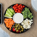 A Fineline black plastic 7-compartment tray with broccoli, baby carrots, tomatoes, and dip.