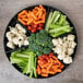 A Fineline black plastic 7-compartment tray filled with carrots and broccoli on a wood surface.