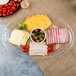 A clear plastic oval catering tray with a stack of cheese, slices of ham, and bowls of olives and cherry tomatoes on a table in a deli.