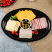 A black Fineline oval cater tray with cheese, ham, olives, and tomatoes.
