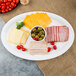 A white plastic oval cater tray with cheese, ham, tomatoes, olives, and peppers.