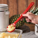 A hand holding a Carlisle red long handle perforated portion spoon filled with pineapple over a bowl of pineapple.