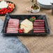 A Fineline black rectangular plastic cater tray with meat, cheese, and other food items on a table with bowls of vegetables and white sauce.