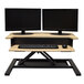 A Luxor white oak adjustable two-tier stand up desktop desk with two computer monitors and a keyboard.