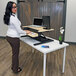 A woman standing at a white oak Luxor two-tier stand up desktop desk with a laptop on the top shelf.