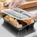A gloved hand putting food into a black Choice 3-compartment plastic container with a lid.