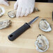 A person in white gloves using a Winco Galveston Style Oyster Opener knife to open oysters on a wood surface.