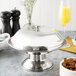 A stainless steel compote dish on a table with food and a glass of orange juice.