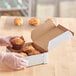 A person in gloves holding a white bakery box filled with muffins.