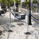 An American Metalcraft polished chrome flat head crowd control stanchion on a stone surface with a blue rope barrier.
