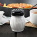 A Tablecraft black ABS plastic perforated salt shaker top on a white table next to a pan of food and a cup of coffee.
